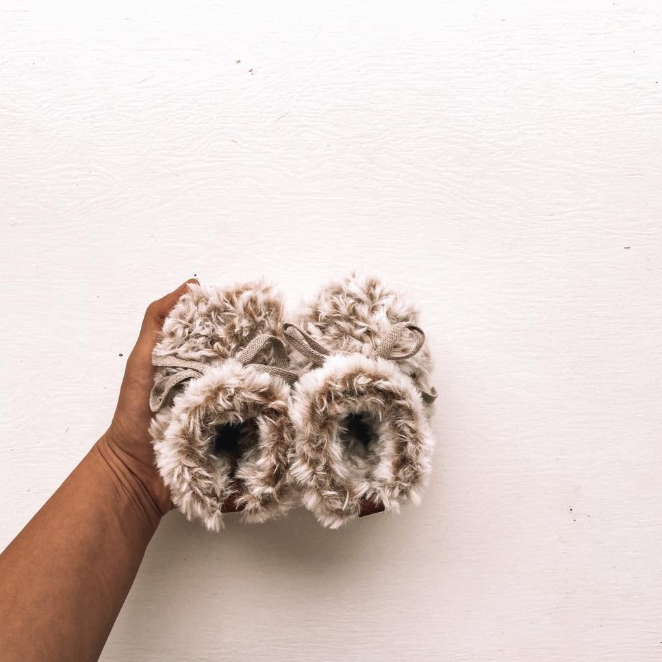 White background with a hand holding the Handknit Teddy Booties by Petit Nordique. Teddy booties are a teddy fur material, with cotton ties.