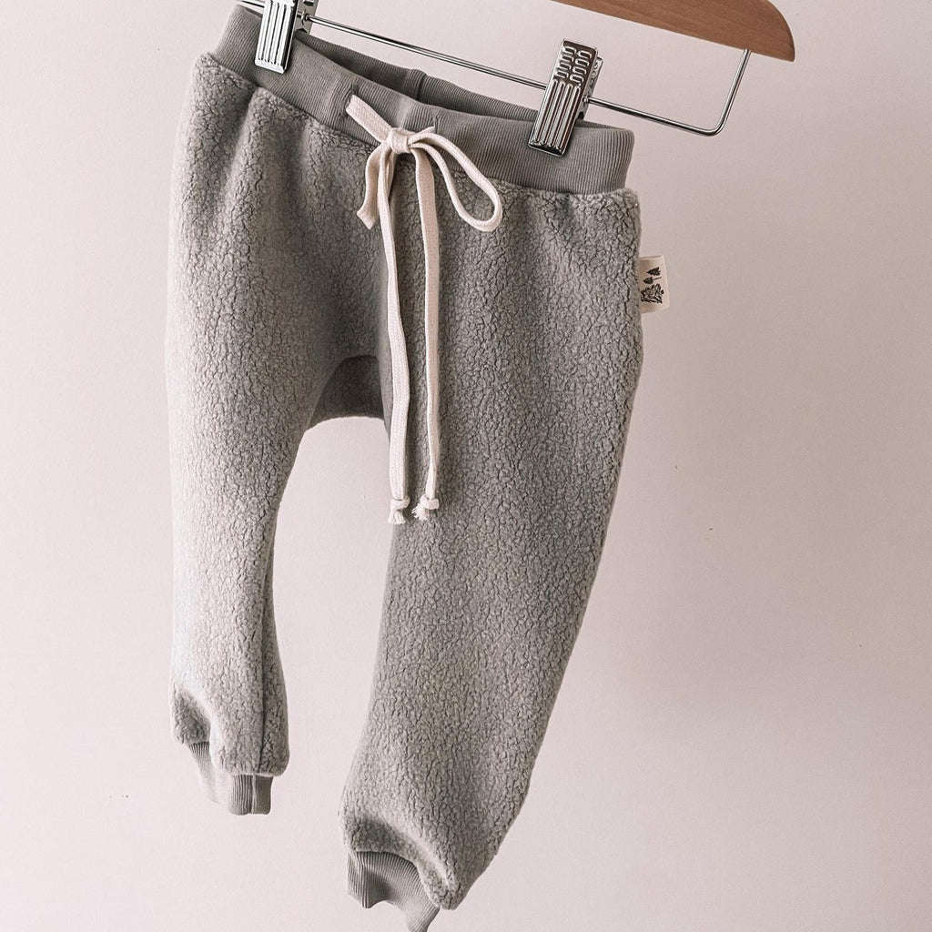 Grey Winter Sky Pant with cream draw string hanging on a metal and wooden pant hanger in front of a white wall. 
