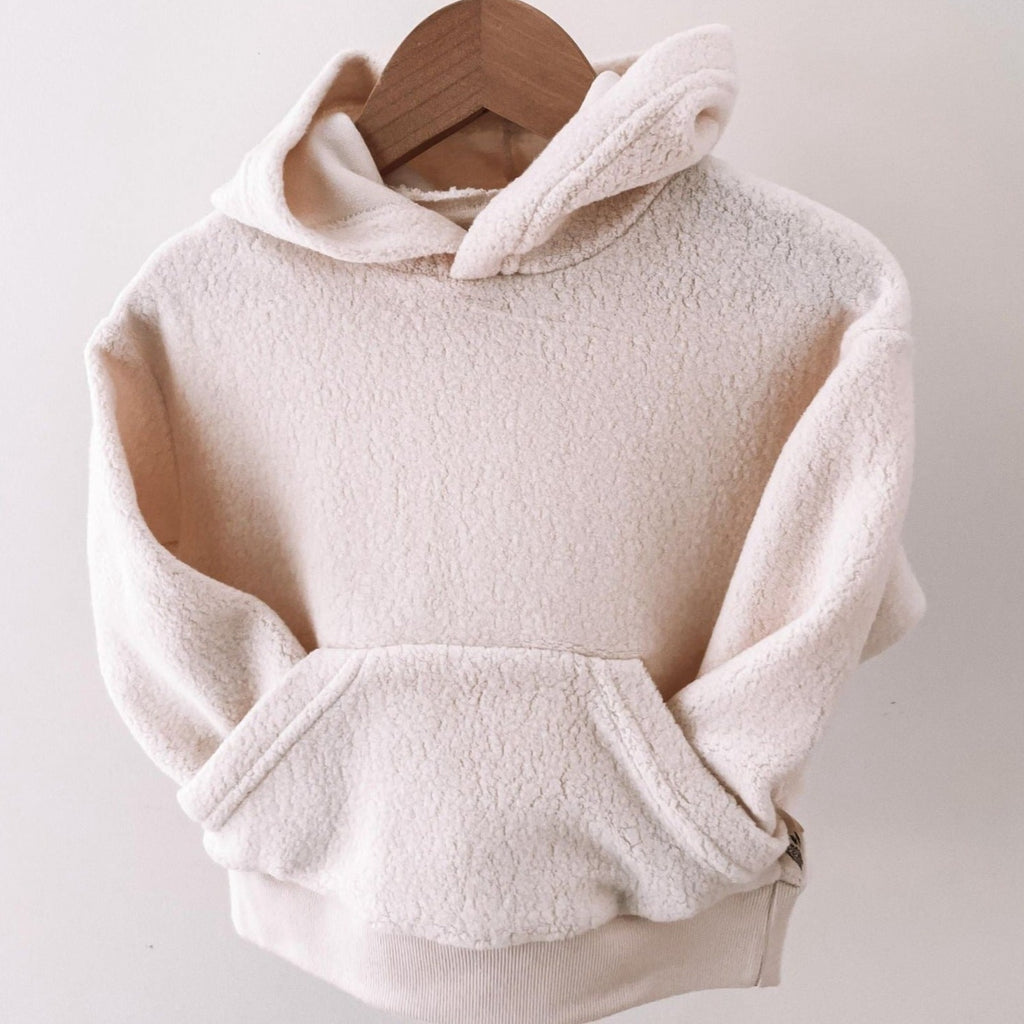 Polar Bear Sherpa Hoodie by Petit Nordique hanging on a wooden hanger in front of a white wall. 