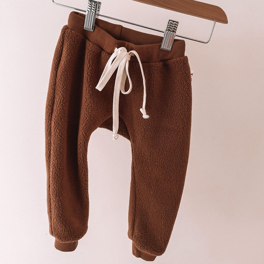 Downtown Brown Sherpa Pant with a cream coloured drawstring, by Petit Nordique, hanging on a wooden and metal pant hanger in front of a white wall. 