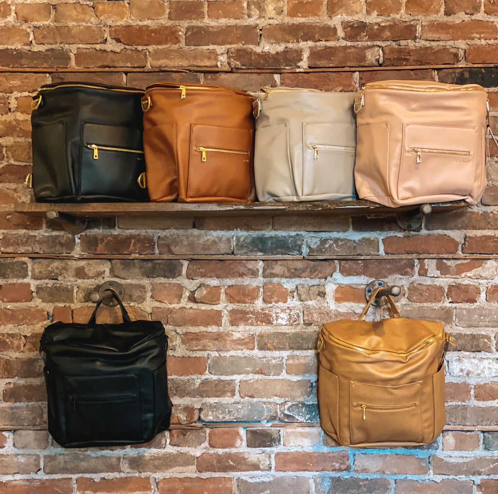 Rustic brick wall with a floating shelf and 4 Original Diaper Bag by Fawn Design on it, and 2 hooks with another 2 Original Diaper Bag by Fawn Design. Showing all of the different colours.