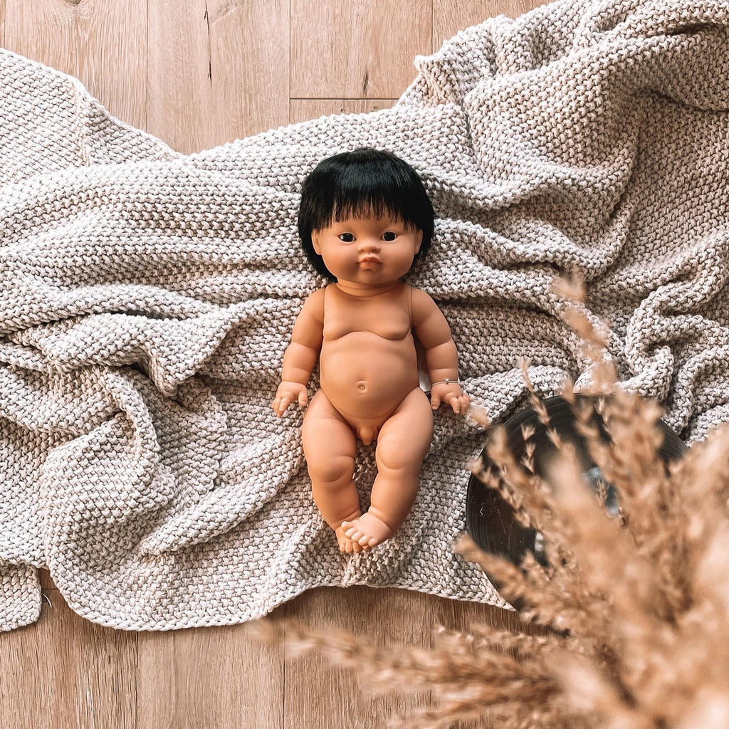 Wood floor with a grey knit blanket, and the Jude Doll by Minikane. Jude is an asian male doll. with short dark hair, and dark eyes.