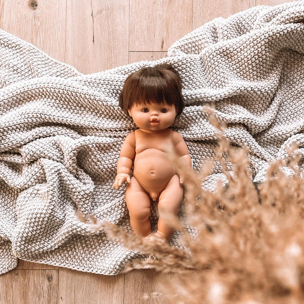 Wood floor with grey knit blanket, pampas grass on the bottom righ corner and the Jules Doll by Minikane laying in the middle. Jules is a caucasian male doll, with short brown hair, and brown eyes.