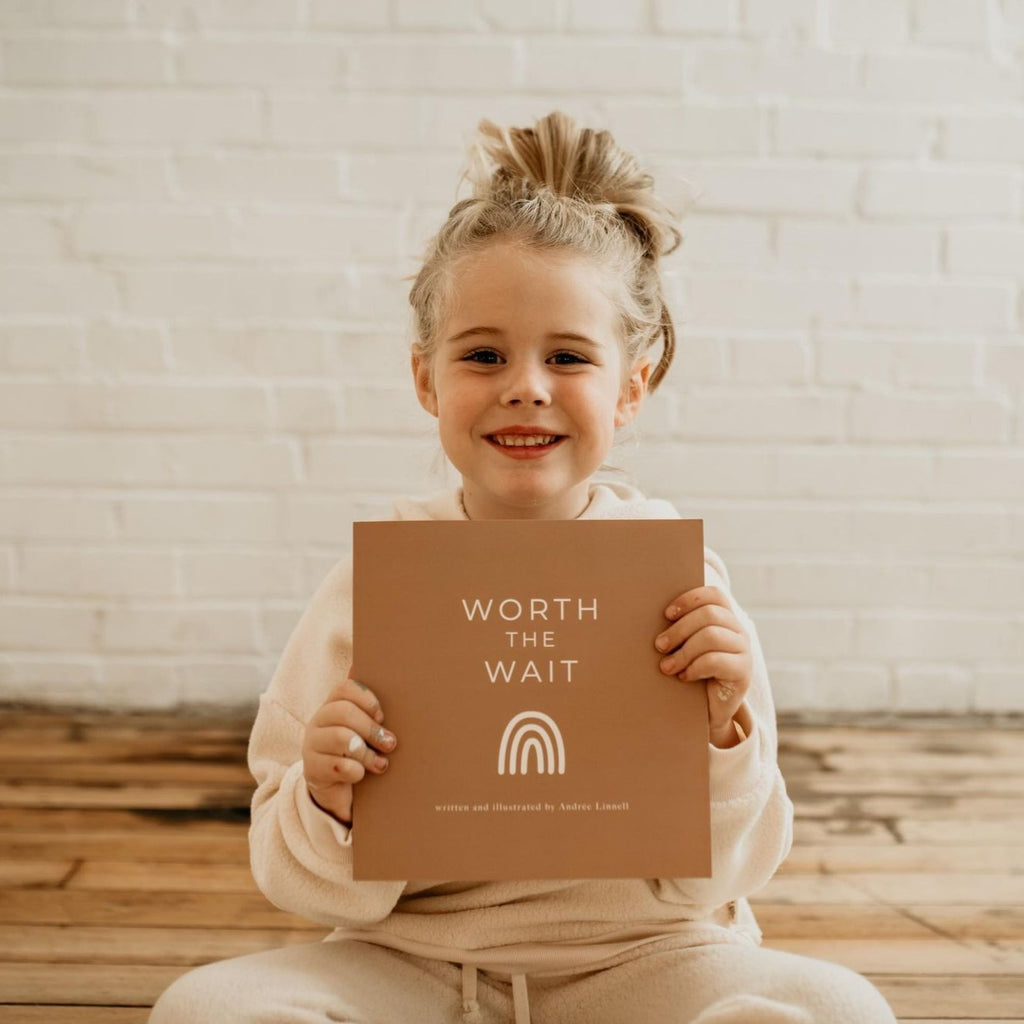 White brick wall with natural wood floors and a little girl sitting, smiling at the camera and holding the 'Worth The Wait' Book | Written & Illustrated by Andrée Linnell. Cover is brown and says "Worth the wait" in white, with a white rainbow.