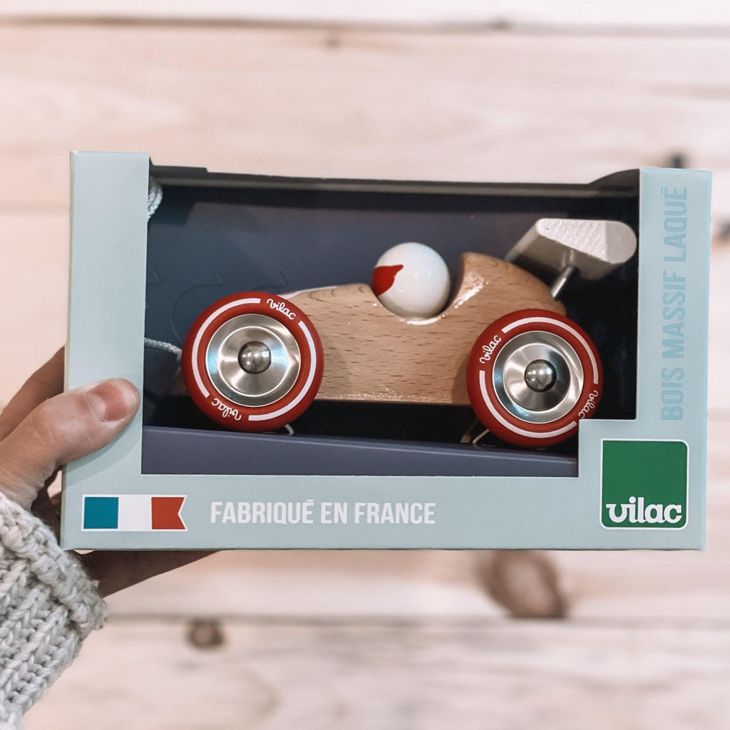 A hand holding p the Pull Along Racing Car in Natural by Vilac. Showing the car in the box, the car is a natural wood colour with red wheels.