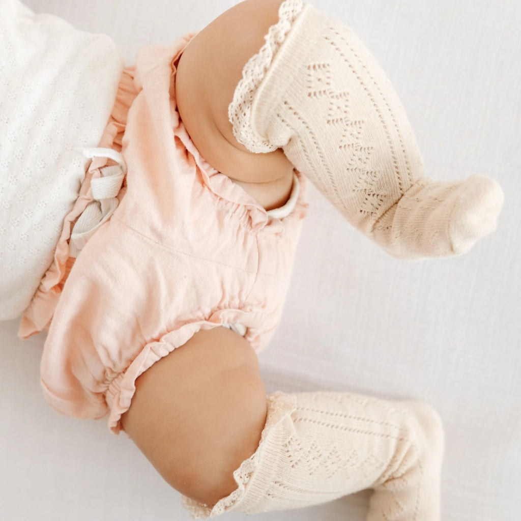 White background with baby laying down in pink bloomers, legs in the air, wearing Fancy Lace Knee High Socks in Vanilla by Little Stocking Co.