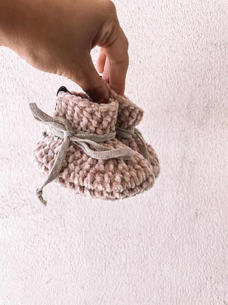 White background with a hand holding the Handknit Velvet Booties in Rose by Petit Nordique. Booties are a soft pink velvet with cotton ties.