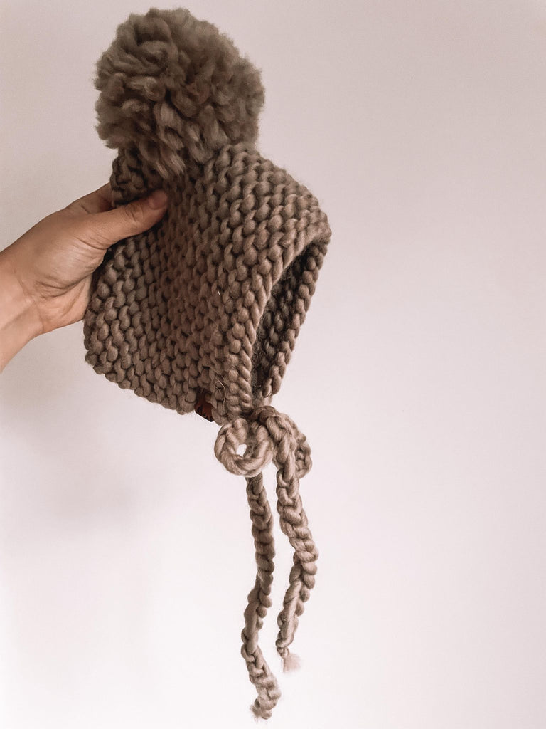White background with a hand holding up The Handknit Bonnet in Taupe by Petit Nordique. Bonnet is a taupe knit, with a matching taupe pompom.