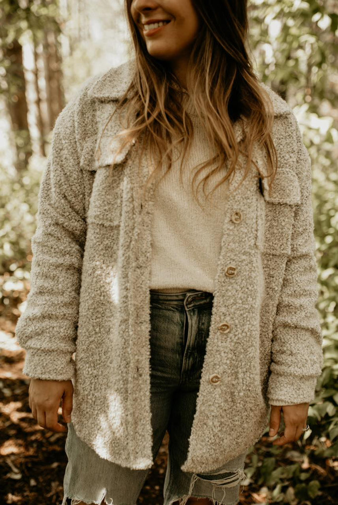 Close up of woman standing, smiling, trees all around her, and she's wearing The Nordique Women's Shacket 2.0 in Oat by Petit Nordique. The shacket is an oat boucle fabric, with buttons down the front, and 2 pockets with button closures on the chest.