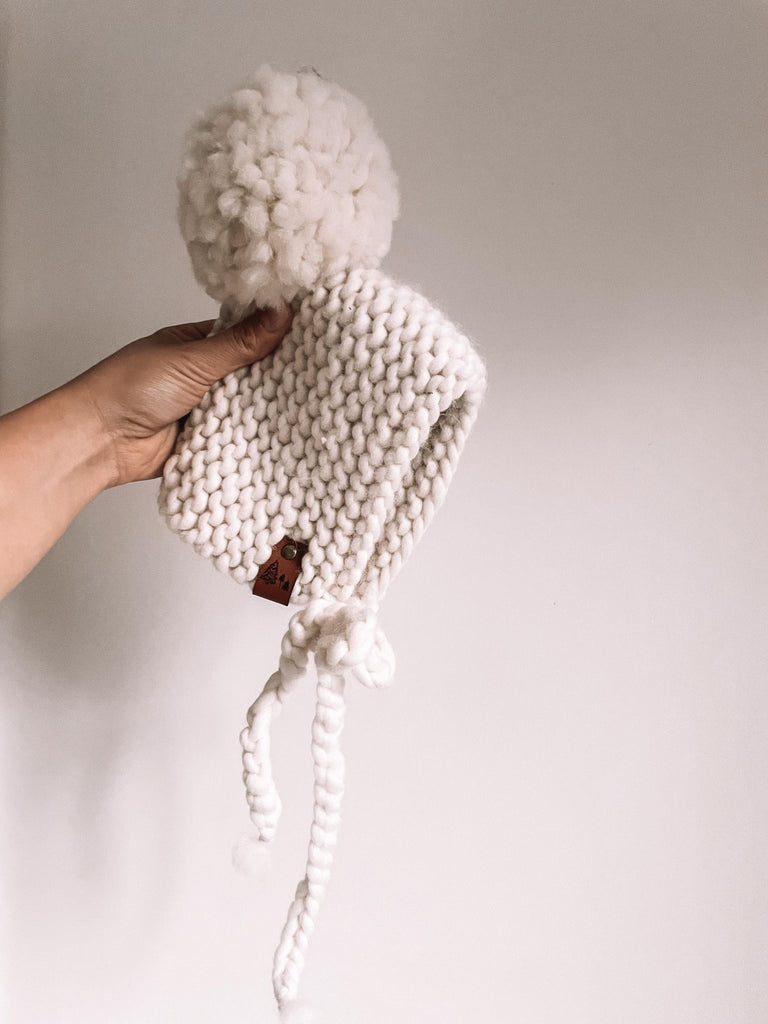 White background with a hand holding The Handknit Bonnet in Natural by Petit Nordique. Bonnet is a creamy natural  yarn, with matching pompom.