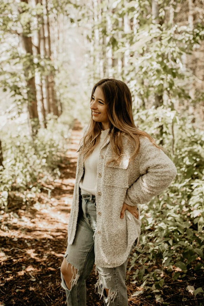 Woman standing, looking to the side, trees are all around her, and she's wearing The Nordique Women's Shacket 2.0 in Oat by Petit Nordique, The shacket is an oat boucle fabric, with buttons down the front and 2 button closure pockets on the chest.