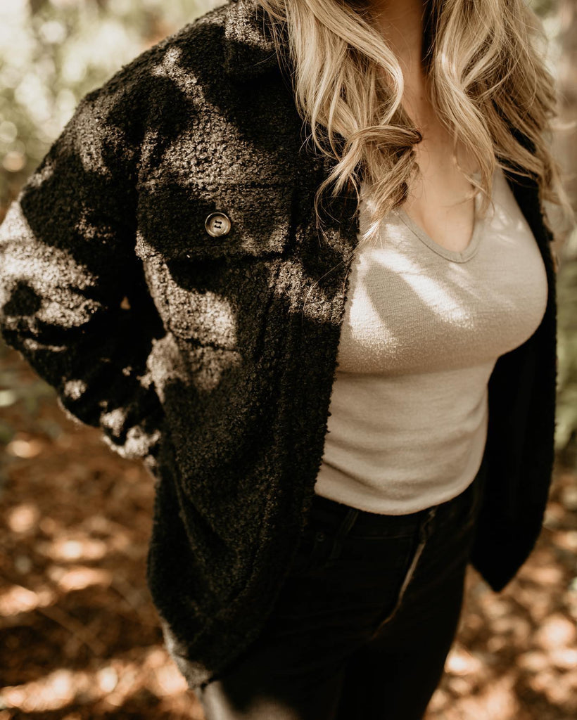 Close up of a woman standing, tree's behind her, and leaves on the ground, wearing The Nordique Women's Shacket 2.0 in Black by Petit Nordique. Showing the button closure on the chest pockets.