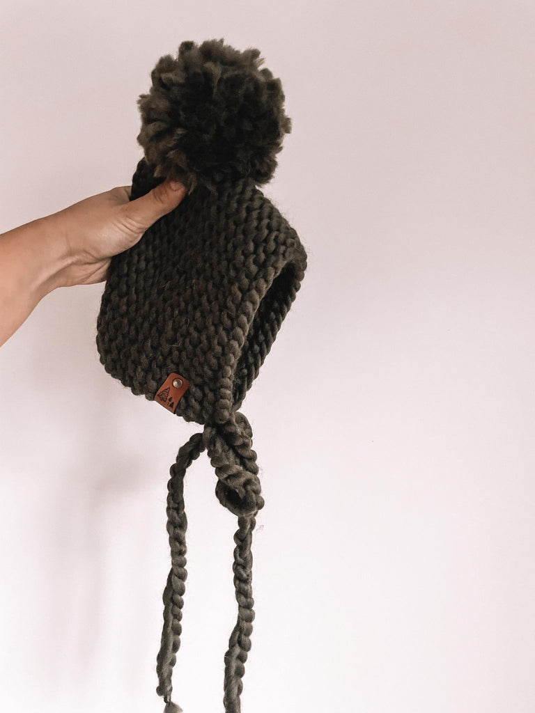 White background with a hand holding The Handknit Bonnet in Olive by Petit Nordique. Bonnet is an olive yarn with a matching pompom.