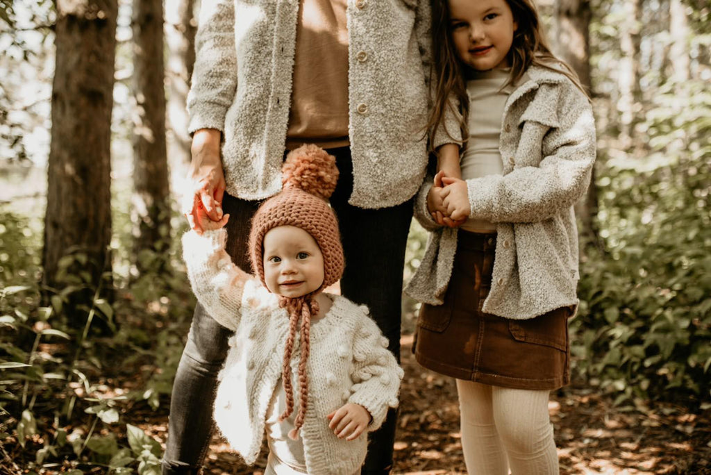 Close up of mom standing with 2 little girls outside in a forest, one of the girls is wearing The Nordique Kids Shacket 2.0 in Oat by Petit Nordique.