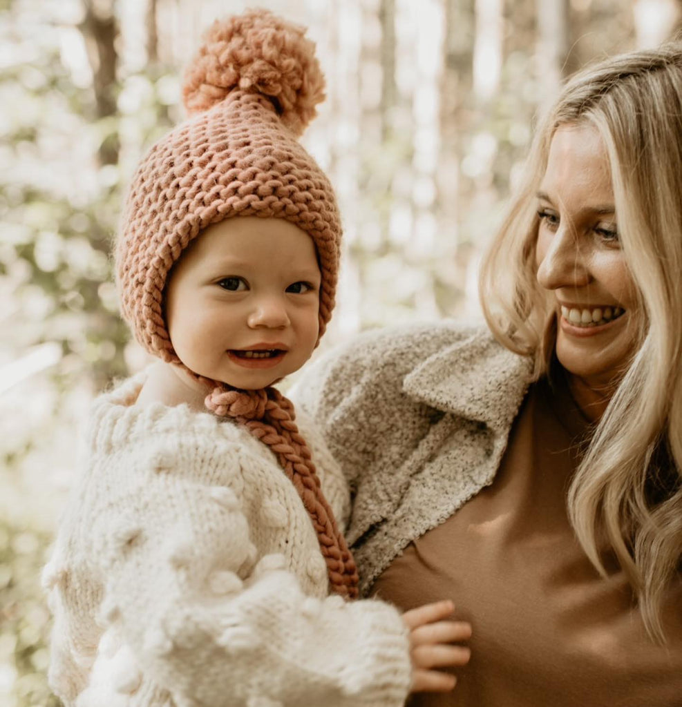 Mama holding a baby girl, trees behind them, she's wearing The Handknit Bonnet in Dusty Rose by Petit Nordique.