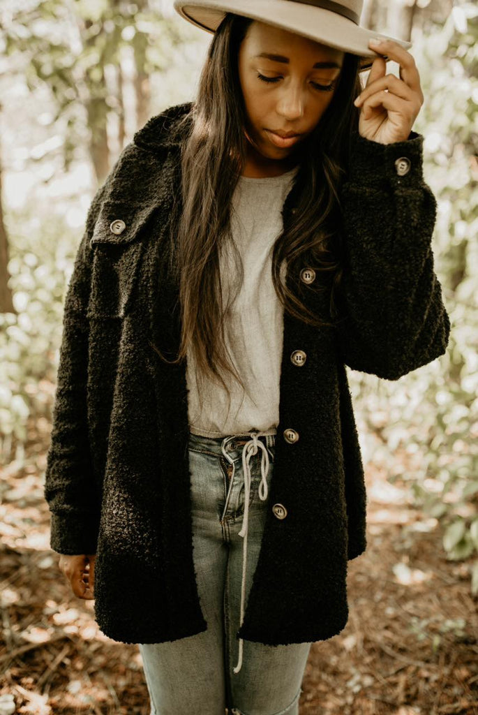 Woman standing, trees behind her, and she's wearing The Nordique Women's Shacket 2.0 in Black by Petit Nordique. This shacket is a black boucle fabric, with buttons down the front and 2 button closure pockets on the chest.