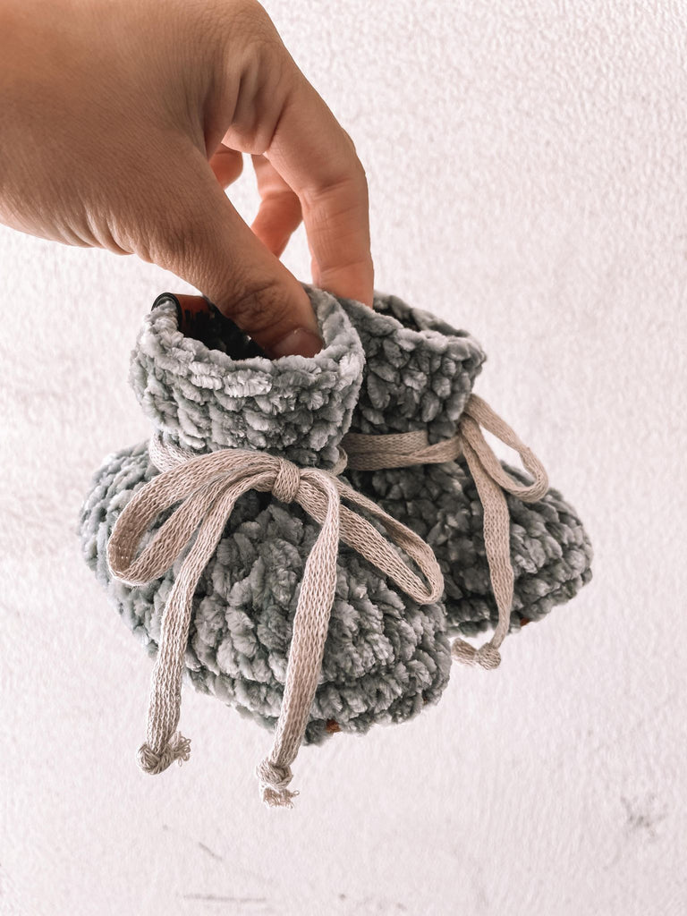White background with a hand holding the Handknit Velvet Booties in Eucalyptus by Petit Nordique. Booties are a green/blue velvet with cotton ties.