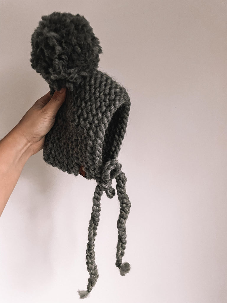 White background with a hand holding The Handknit Bonnet in Grey by Petit Nordique. Bonnet is a dark charcoal, with a matching pompom.