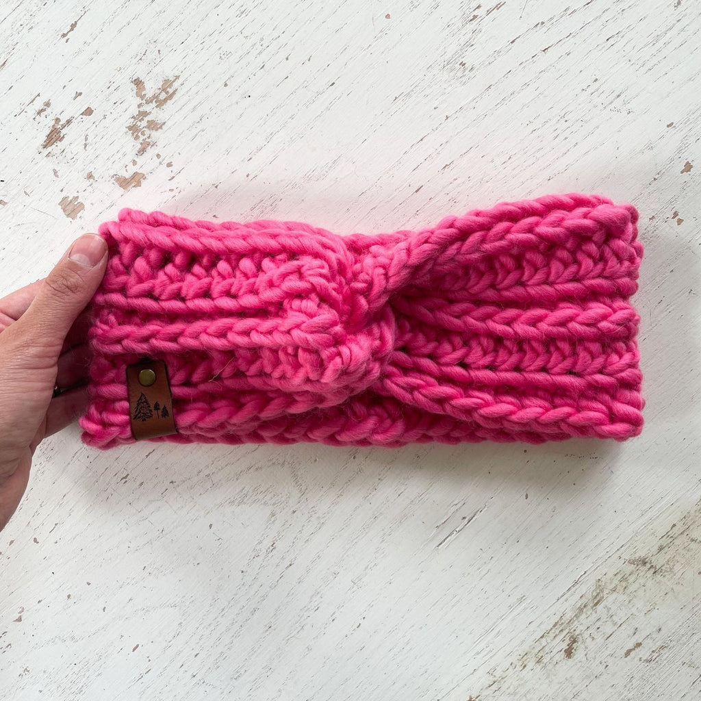 White washed wood background with a hand holding The Chunky Twist Headband in Magenta by Petit Nordique. Headband is a bright pink wool with a small leather tag with trees on it.