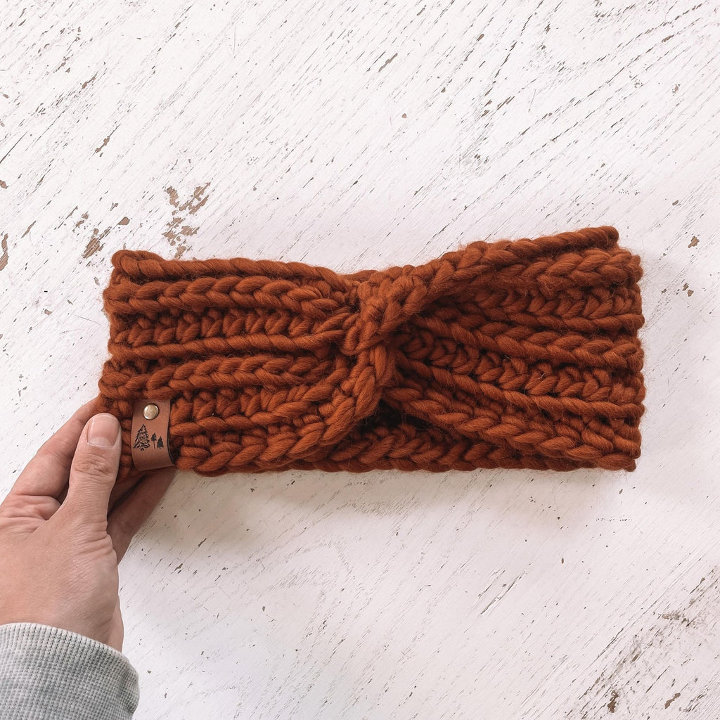 White washed wood background with a hand holding The Chunky Twist Headband in Rust by Petit Nordique. Headband is a rust colour, with a small leather tag with trees on it.
