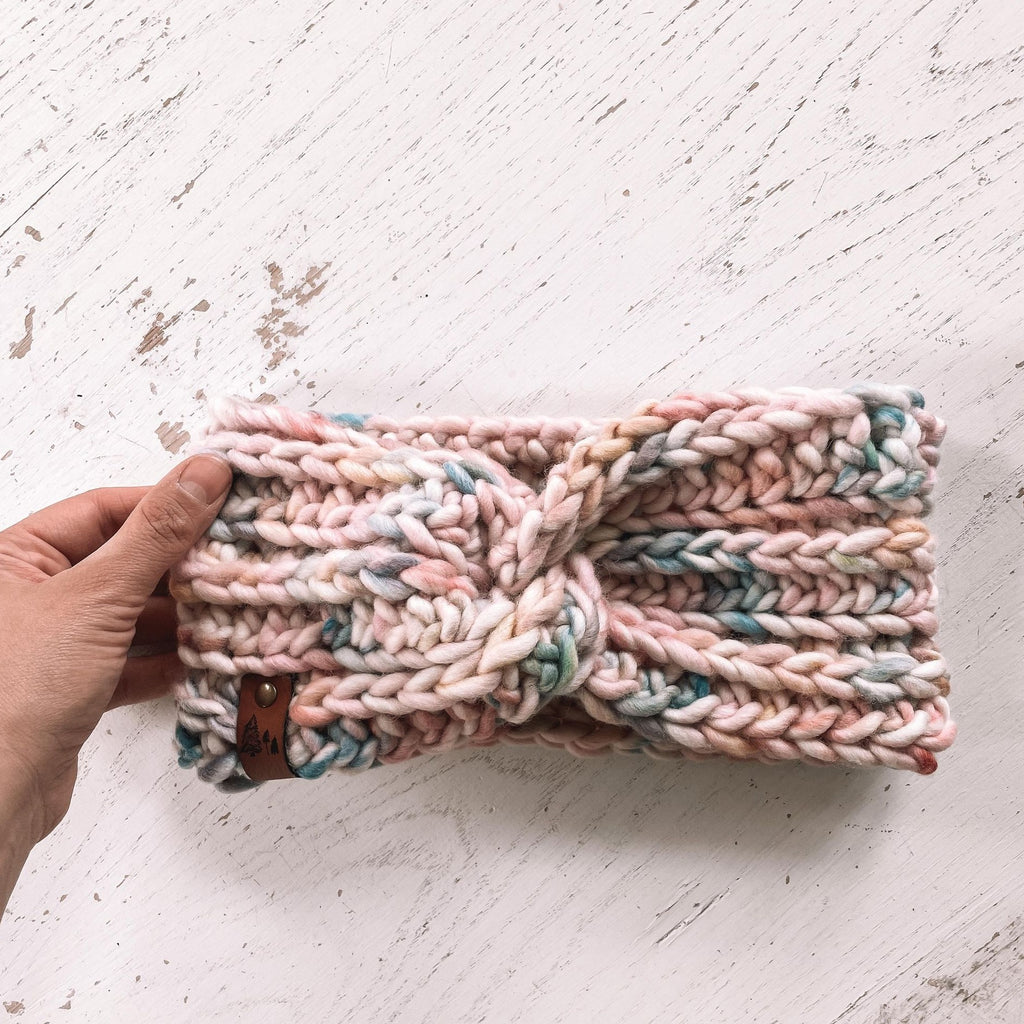 White washed wood background with a hand out, holding onto The Chunky Twist Headband in Coralie by Petit Nordique. Headband is a chunky multicoloured wool with a small leather tag with trees on it.