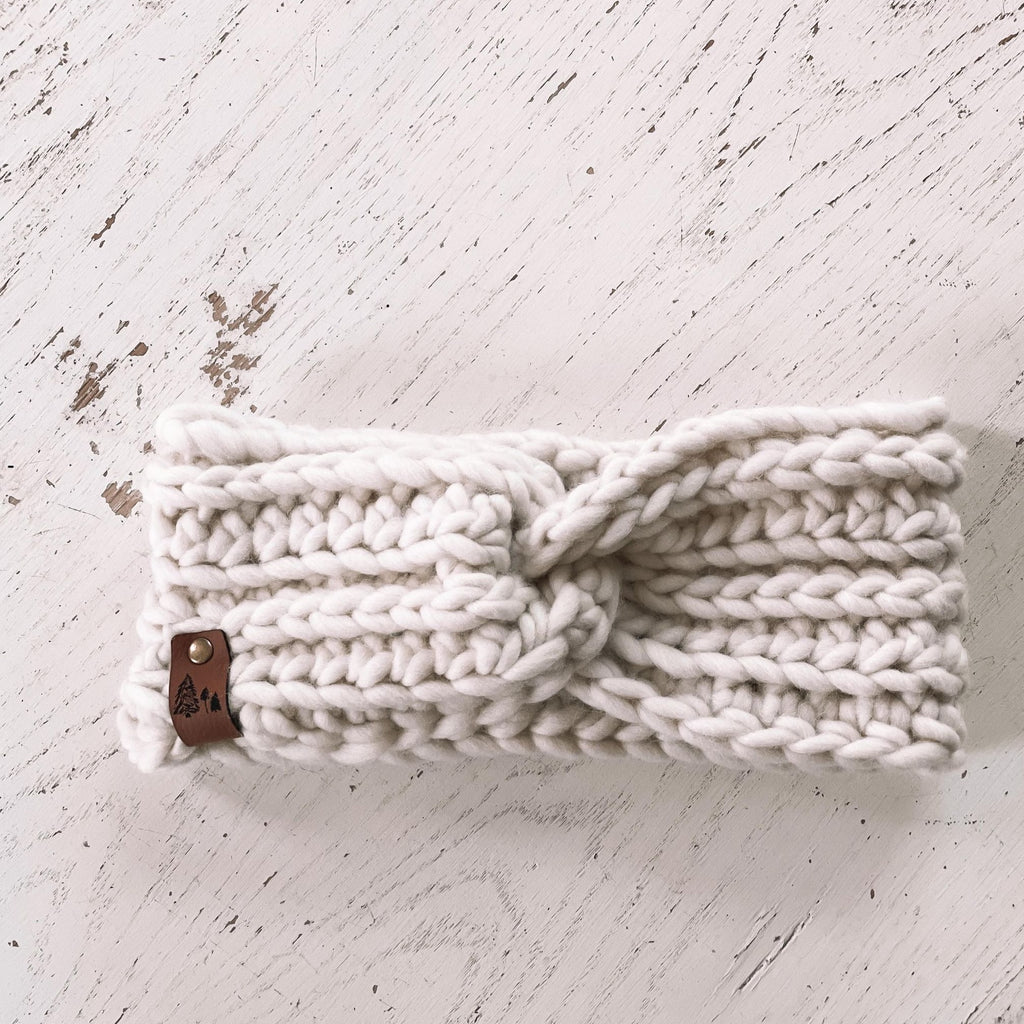 White washed wood with The Chunky Twist Headband by Petit Nordique. Headband is a white/cream wool with a small leather tag with trees on it.