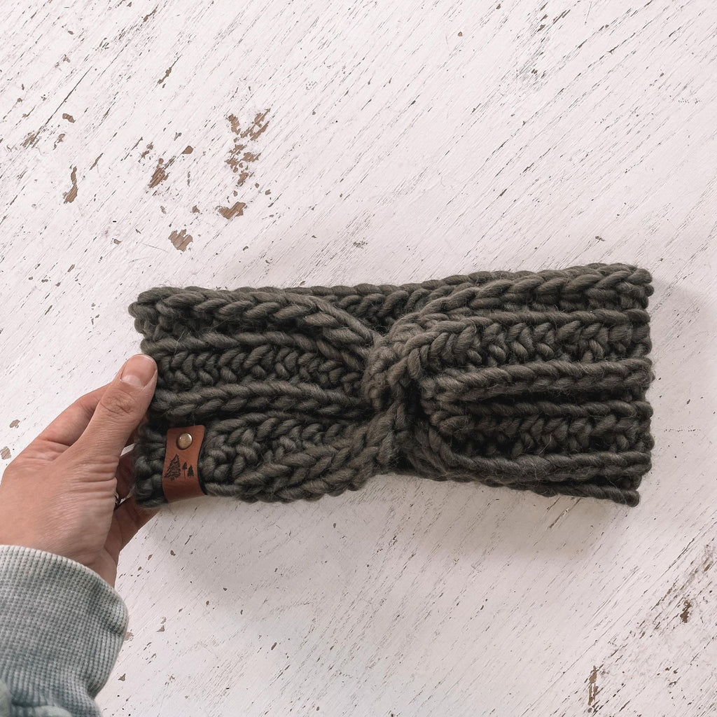 White washed wood background with a hand holding The Chunky Twist Headband in Olive by Petit Nordiquee. Headband is an olive wool, with a small leather tag with trees on it.