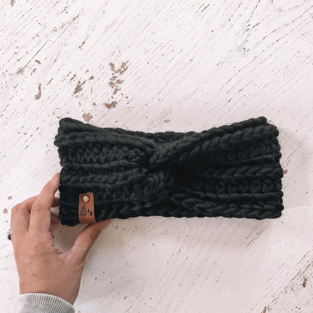 White washed wood background with a hand out, holding The Chunky Twist Headband in Black by Petit Nordique. Headband is a chunky black wool, with a small leather tag with trees on it.