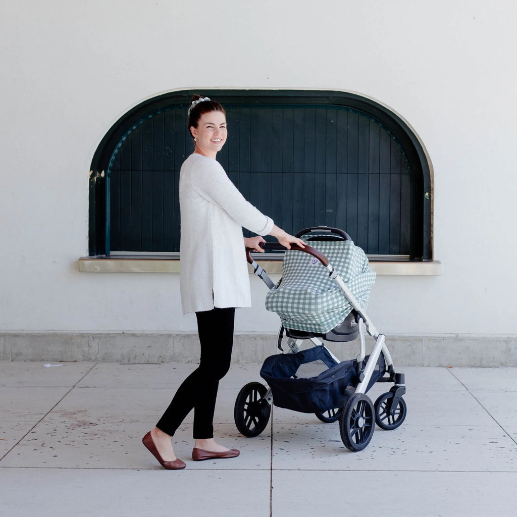 White wall with a black arch window, and a mama pushing her baby in a stroller with The OVer Cover in Paxton by The OVer Company on her car seat. This cover is a sage/blue & white gingham, with a white tag on the front that says "OV' in black, with black greenery.