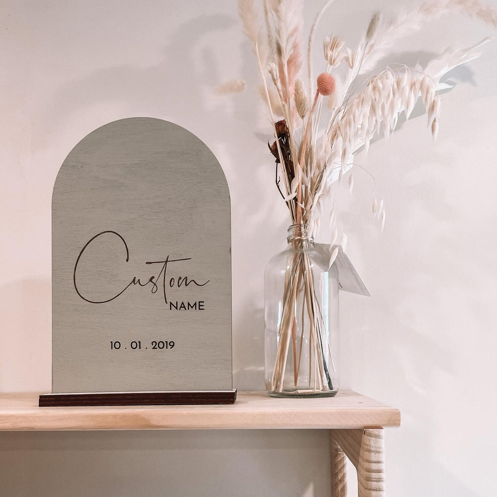 White wall with a natural wood shelf, and the Personalized Name Arch in Dune by Petit Nordique beside a jar of pampas grass. Name arch is an arch, in a sage/grey colour with custom writing on a wooden base.