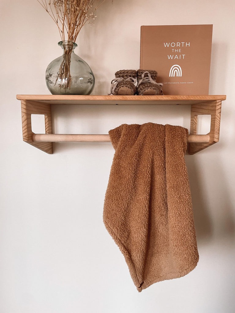 Petit Nordique's Sherpa Blanket in teddy hanging on a wooden rack, with a vase with some grass, Petit Nordique booties in Taupe and a copy of Worth The Wait by Andree Linnell, against a white wall. 