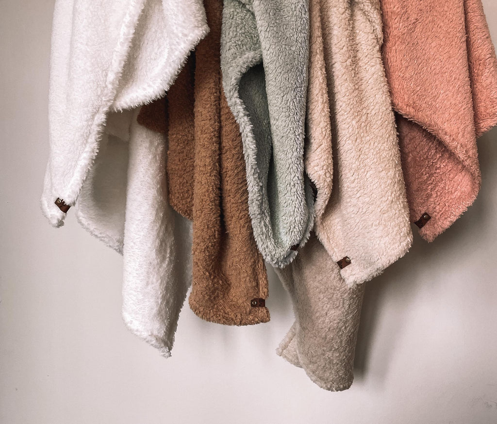 Petit Nordique's new Sherpa Blankets. Pictured left to right in polar bear, teddy, sage, fawn and blush with brown leather tags, against a white wall. 