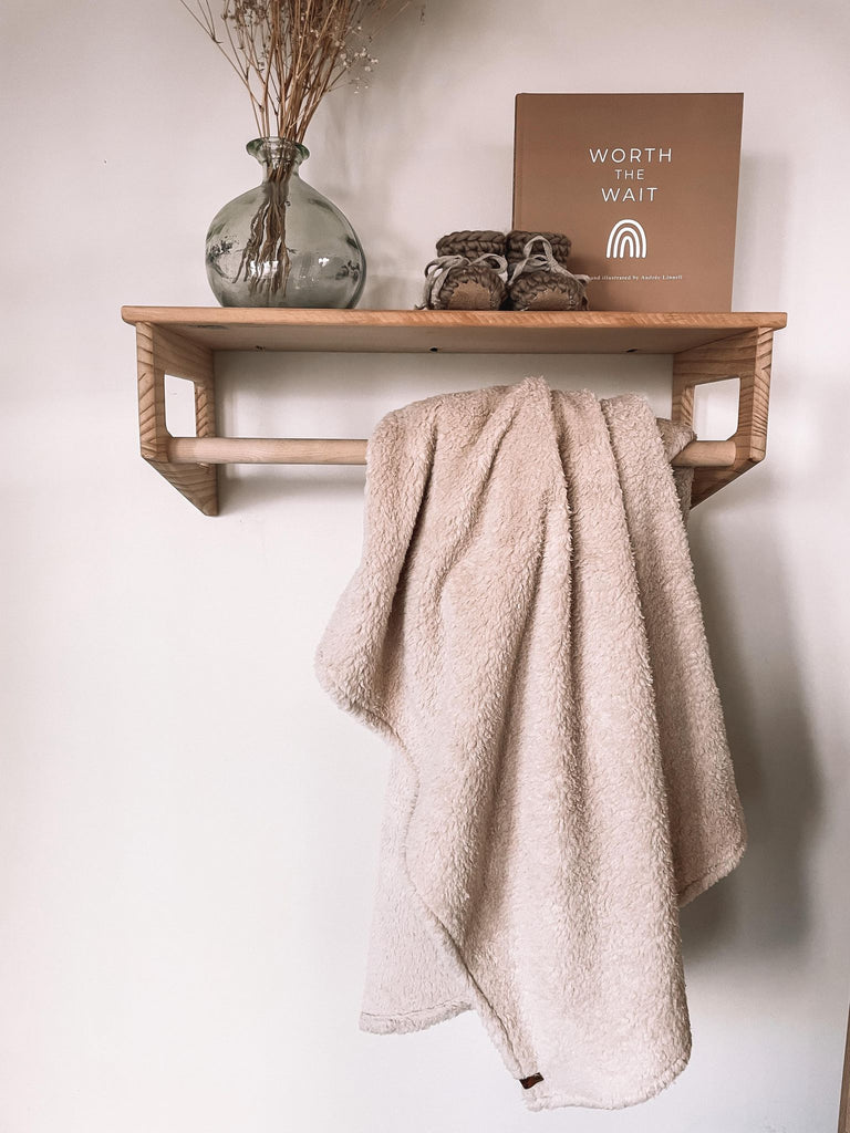 Petit Nordique's Sherpa Blanket in fawn hanging on a wooden rack, with a vase with some grass, Petit Nordique booties in Taupe and a copy of Worth The Wait by Andree Linnell, against a white wall. 