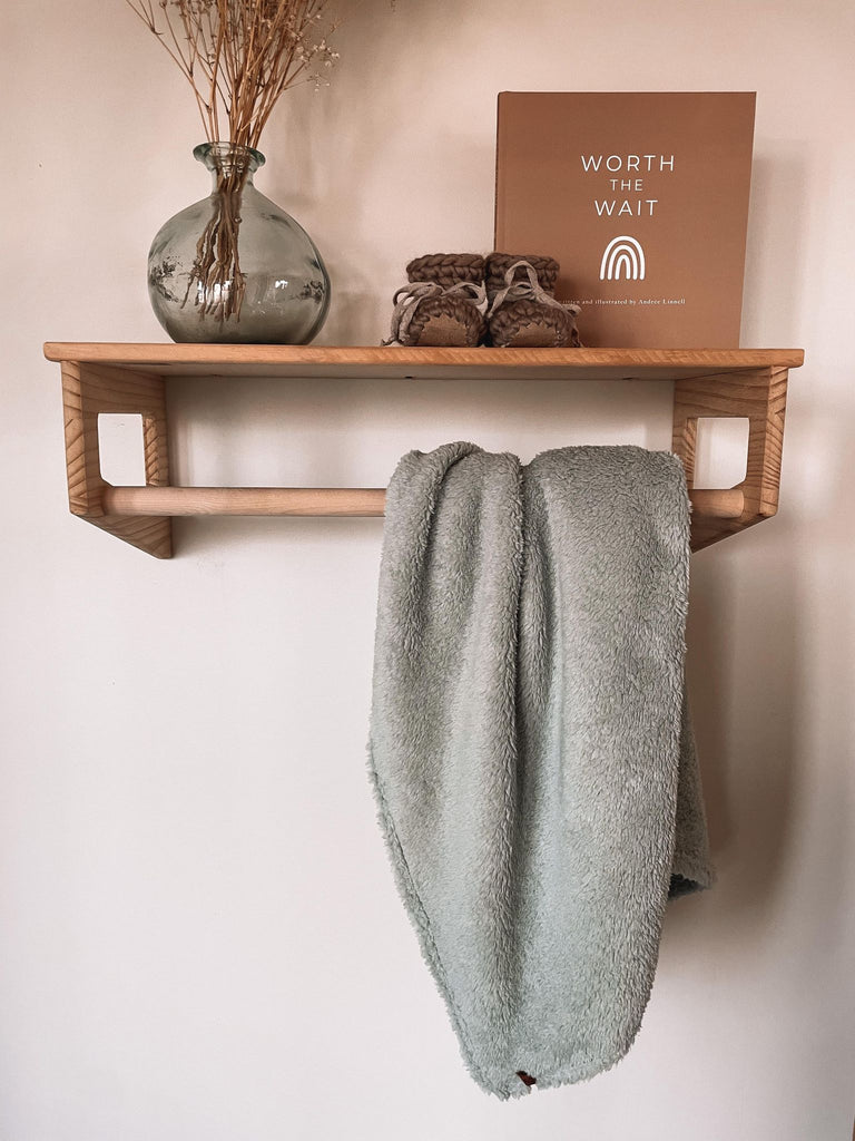 Petit Nordique's Sherpa Blanket in sage hanging on a wooden rack, with a vase with some grass, Petit Nordique booties in Taupe and a copy of Worth The Wait by Andree Linnell, against a white wall. 