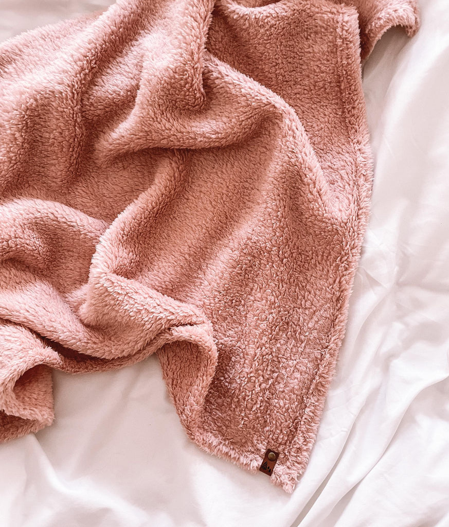 Petit Nordique's blush sherpa blanket spread out on a white a bed sheet. 