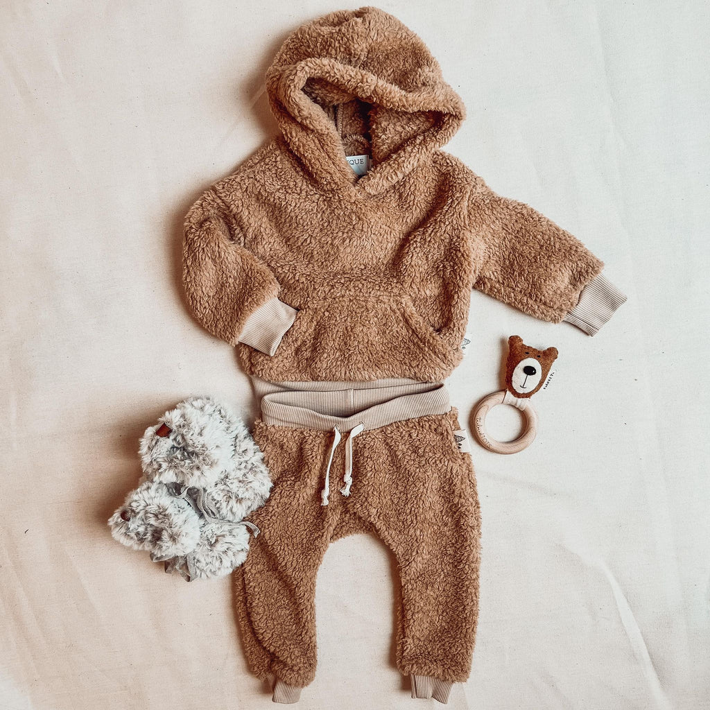 Teddy Sherpa hoodie and pant set by Petit Nordique, with faux fur booties by Petit Nordique as well. A bear rattle beside it on a cream surface. 