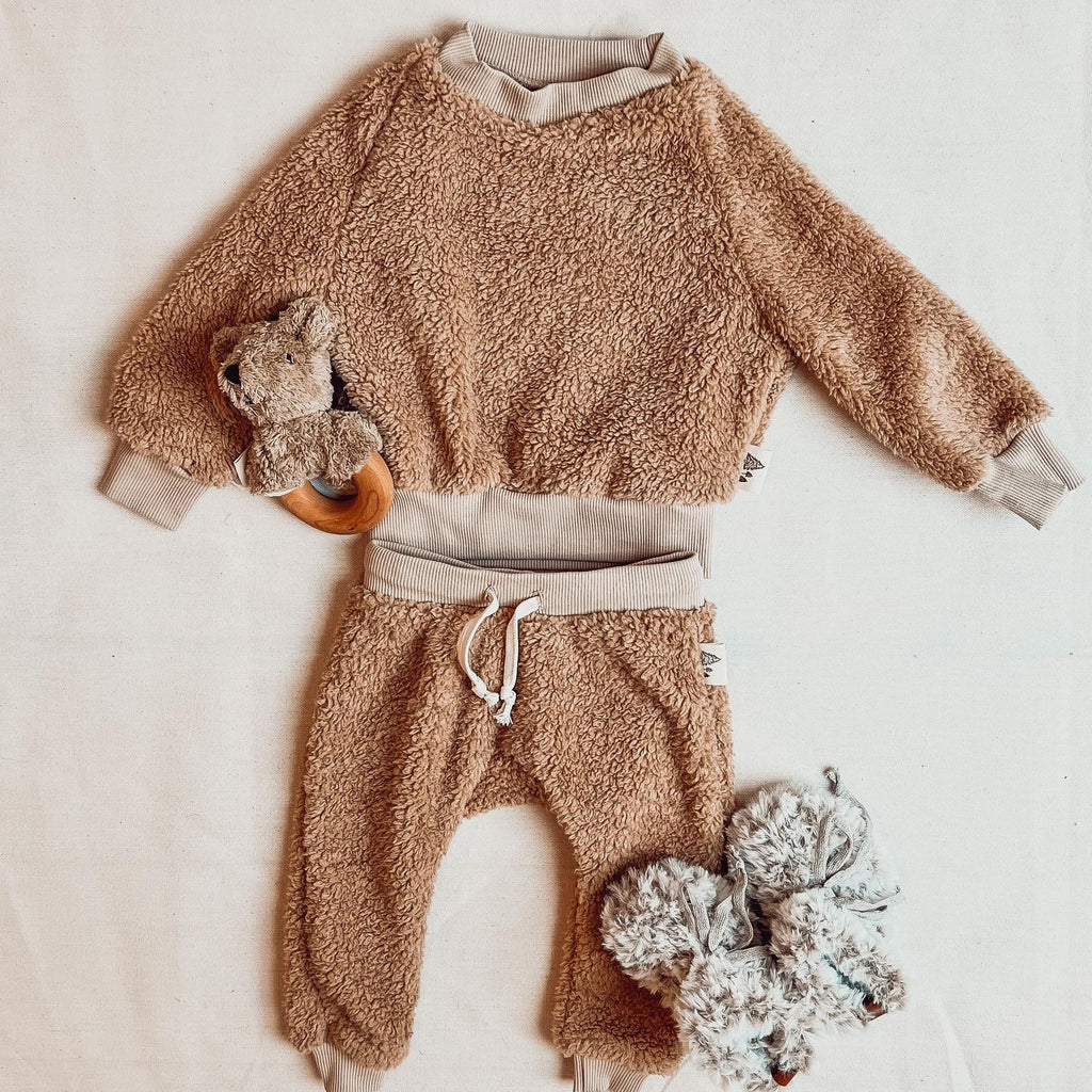 Teddy Sherpa crewneck and pant set by Petit Nordique, with faux fur booties by Petit Nordique as well. A bear rattle beside it on a cream surface. 