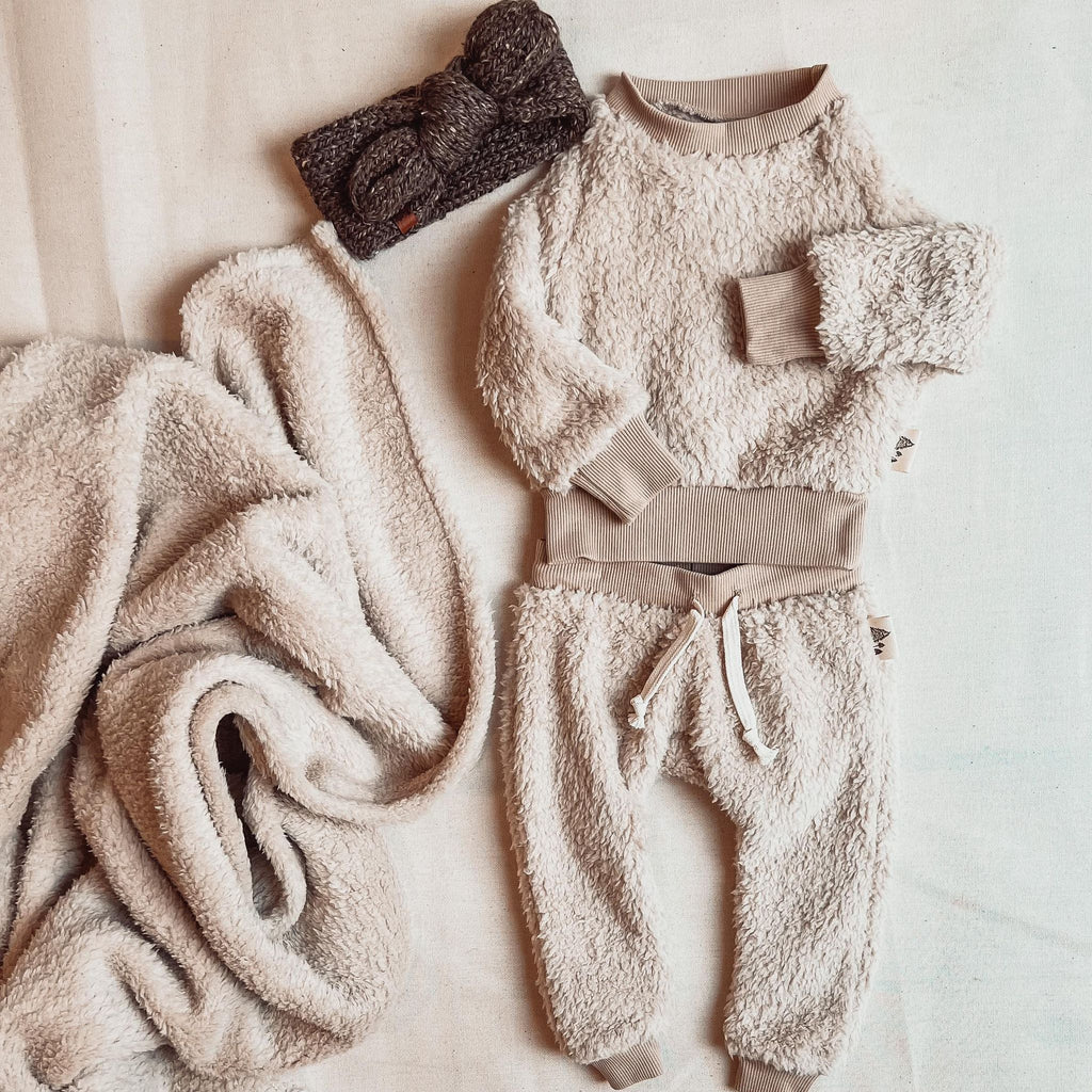 Fawn crew neck sherpa and pants by Petit Nordique, with a brown tweed knot bow by Petit Nordique as well, with a sherpa Petit Nordique blanket beside it. Laid on a cream surface. 