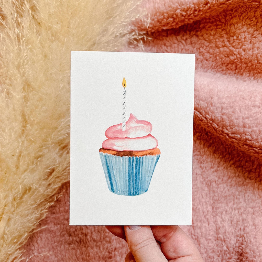 Pink Cupcake card by PARKES & bash being held up by a hand with pampas grass in the background and a pink sherpa blanket by Petit Nordique. 