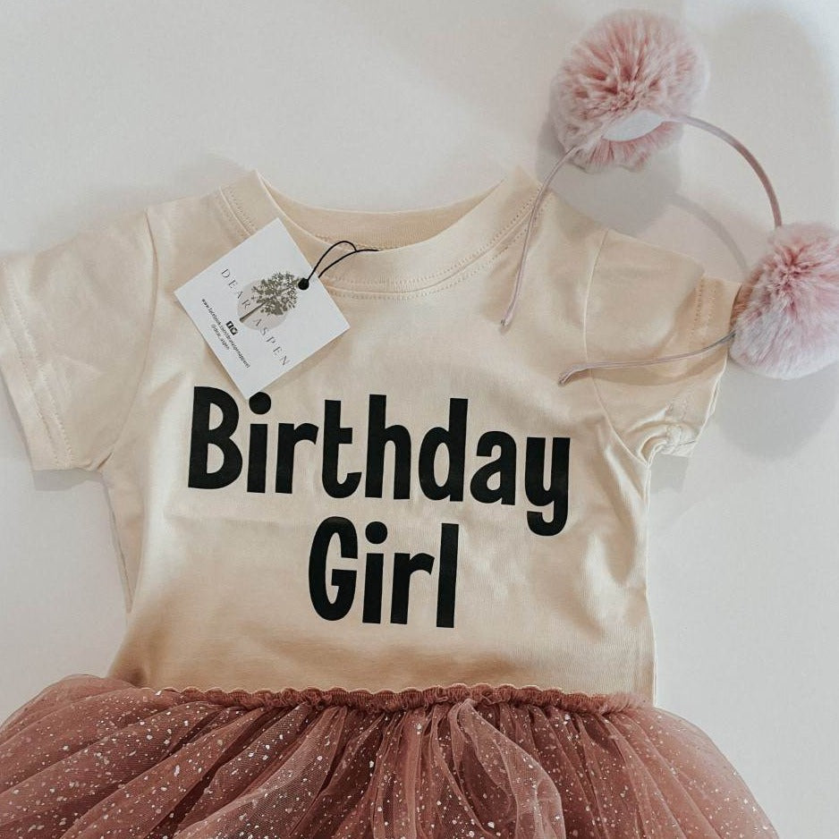 Birthday Boy and Birthday Girl Tees by Dear Aspen paired with a pink tutu and pink pompom headband, laid on a flat white surface. 