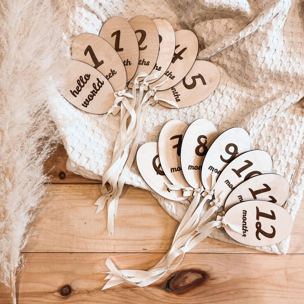 hello world | monthly milestones balloon set by Petit Nordique wooden monthly milestone discs for baby announcements photos all laid out attached with cotton string on wooden table with waffle blanket and pampass grass