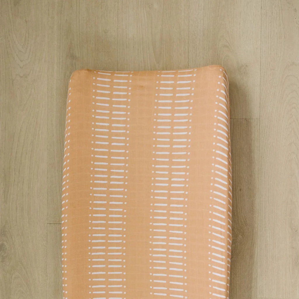 Light wood floor with a change pad and a Sahara Changing Pad Cover by Mebie Baby on it. Cover is pale orange with lines.