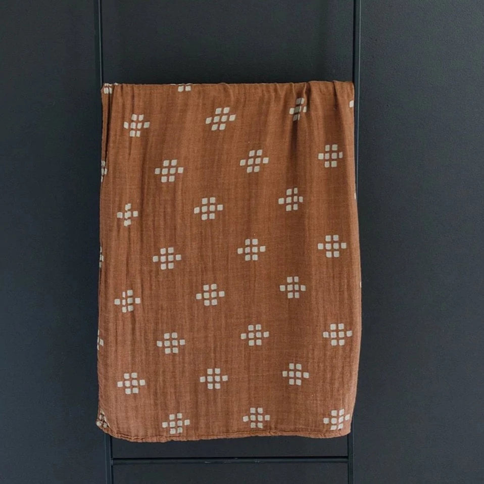 Dark background with a black metal blanket ladder, and a Chestnut Textiles Muslin Swaddle by Mebie Baby. Swaddle is dark brown with cream square pattern all over.