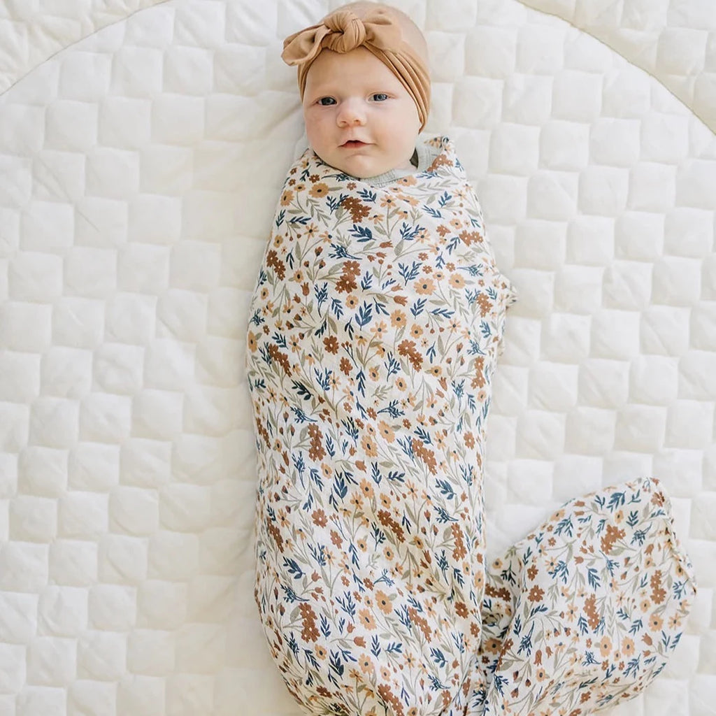Overhead view of baby laying on white, wrapped up in a Harvest Floral Muslin Swaddle by Mebie Baby. Swaddle is white/cream with a close pattern of flowers in rust, blue and beige all over.