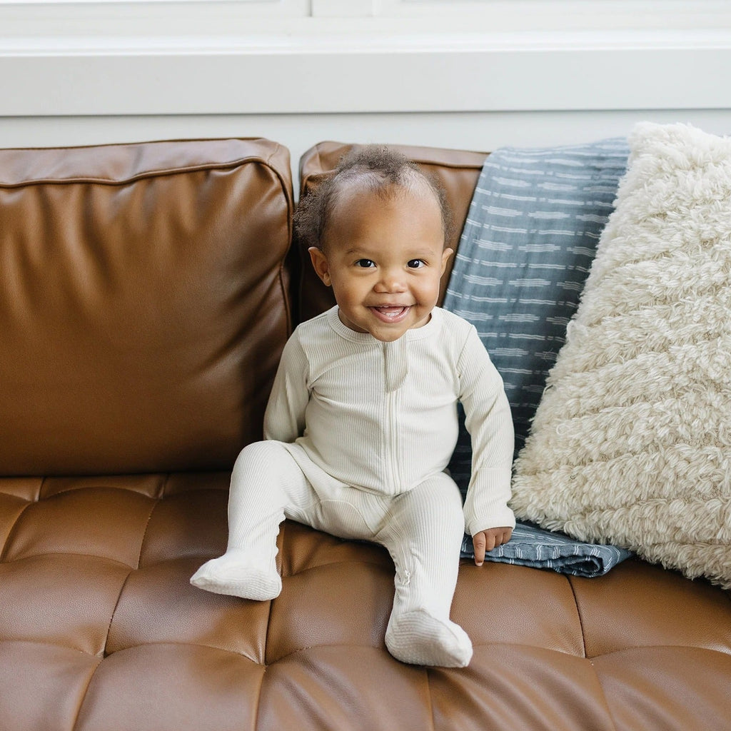 Baby sitting on a brown leather couch, wearing the Vanilla Organic Cotton Footed Zipper One-Piece by Mebie Baby.