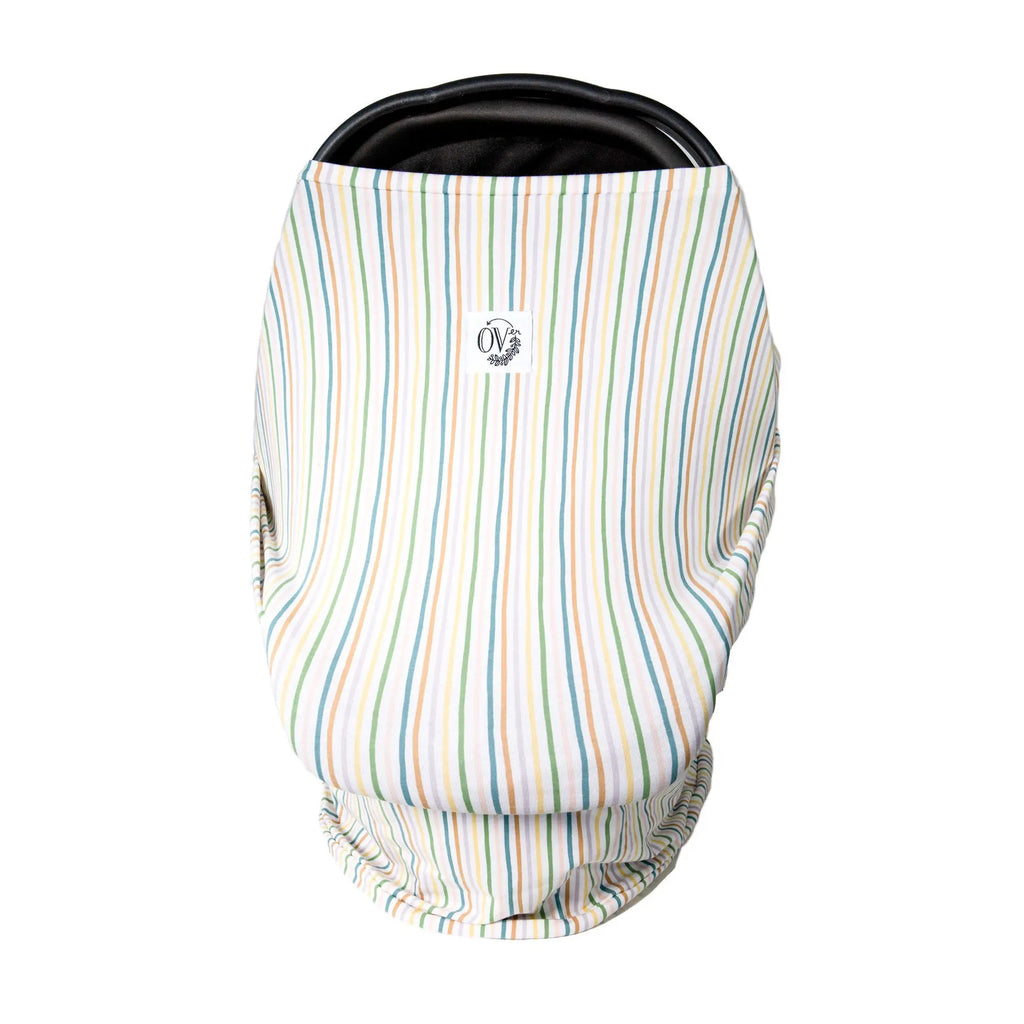 White background with a carseat and The Over Cover in Promise by The Over Company is on her car seat. This cover is white with multi coloured vertical lines, with a white tag on the front with black font that says “OV” with greenery.