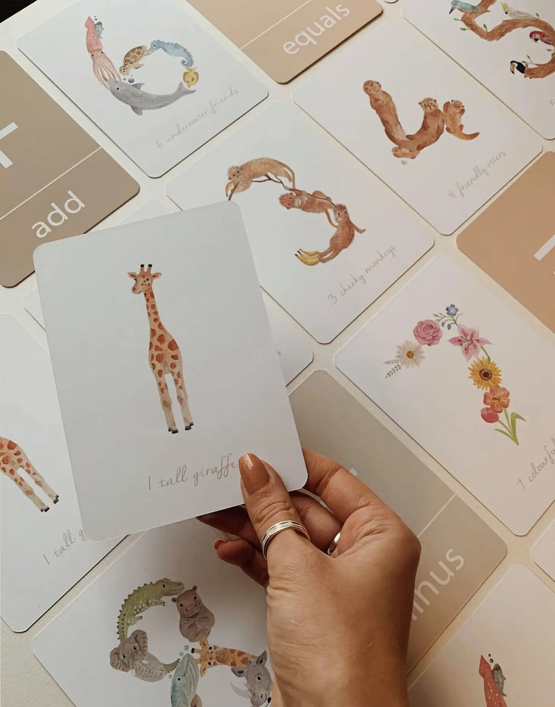 Overhead view of Number Flash Cards by Little Roglets laid out. Hand is reaching out holding a white card with a giraffe in the middle and the words "1 tall giraffe" on the bottom.