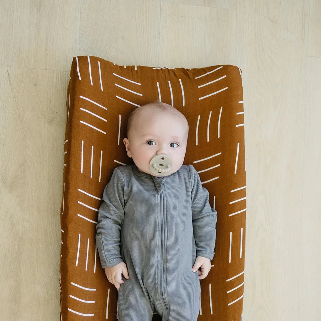 Light wood floor with overhead view of baby laying on change pad with a Mustard Mudcloth Changing Pad Cover by Mebie Baby on it. Changing pad cover is mustard with white lines, and fits snug.
