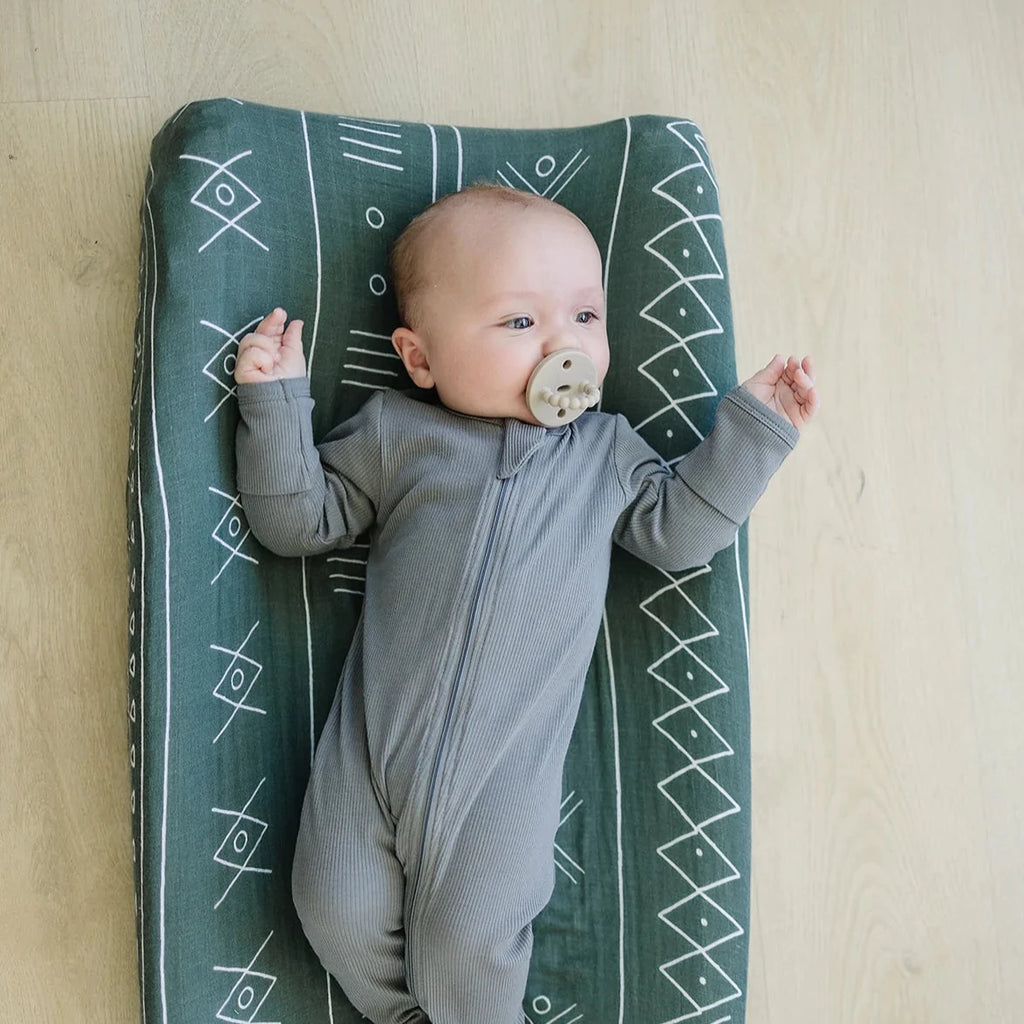 Wood floor background with baby laying on changing pad with an Alpine Changing Pad Cover by Mebie Baby. Changing pad cover fits snug, it's green with an aztec print.