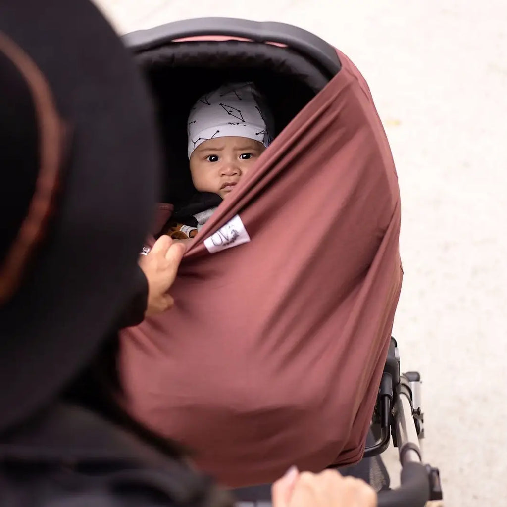 Close up overhead view of a mama pushing her baby in a stroller, with The OVer Cover in Lennon by The OVer Company covering the car seat, and the mom is pulling the front down. The colour is a burgundy red, with a white tag and black text that says "OV" with black greenery.